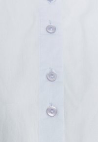 Chemise bleu clair à manches longues avec une texture lisse, comprenant six boutons ronds et brillants ainsi que des détails de patte de boutonnage cousue.