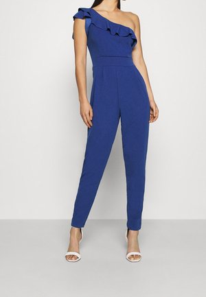 Woman wearing a royal blue one-shoulder jumpsuit with ruffle detail and tapered legs, paired with white open-toe high-heeled sandals.