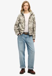 Veste légère à motif camouflage dans des tons gris, dotée d'une fermeture éclair sur le devant, d'un col montant et de poches latérales, portée avec un jean en denim ample et des bottes.