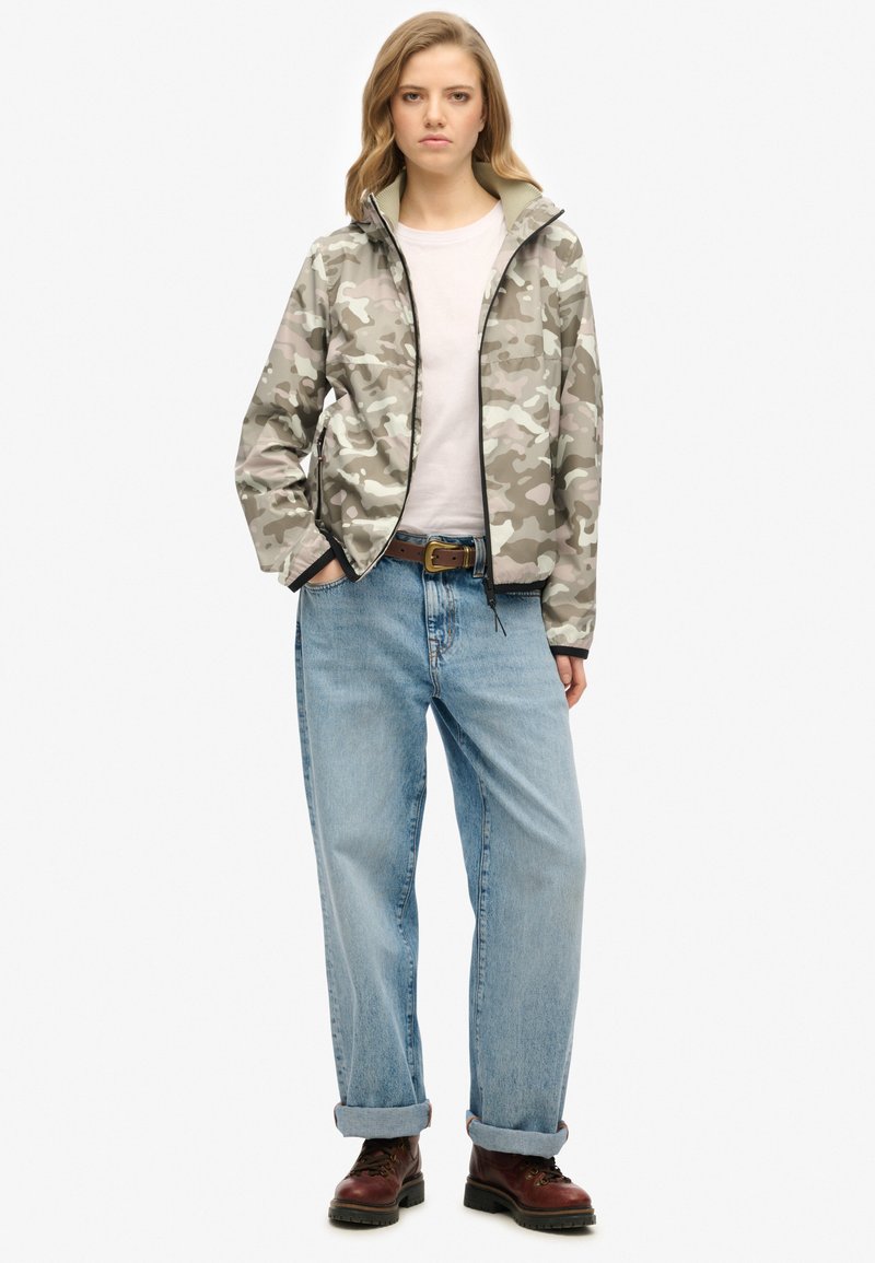 Veste légère à motif camouflage dans des tons gris, dotée d'une fermeture éclair sur le devant, d'un col montant et de poches latérales, portée avec un jean en denim ample et des bottes.