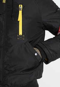 Veste matelassée noire avec fermeture éclair jaune, logo étoile brodé sur la poitrine, patch rouge sur la manche et main dans la poche à pression.
