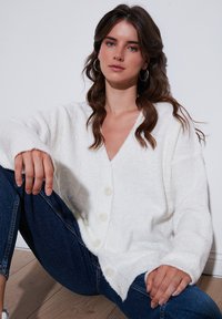 Cardigan alb, pufos, cu decolteu în V și nasturi mari, având manșete și tiv cu dungi. Purtați peste blugi albaștri, așezat pe o podea din lemn.