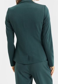 Tyrkysový na míru šitý blazer se štíhlým střihem, strukturovanými rameny a hladkým materiálem. Obsahuje jeden rozparek na zadní straně a dlouhé úzké rukávy.