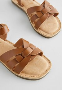 Sandali in pelle marrone caratterizzati da ampii cinturini incrociati e suola piatta. La soletta ha una consistenza morbida, con cuciture lungo i bordi.