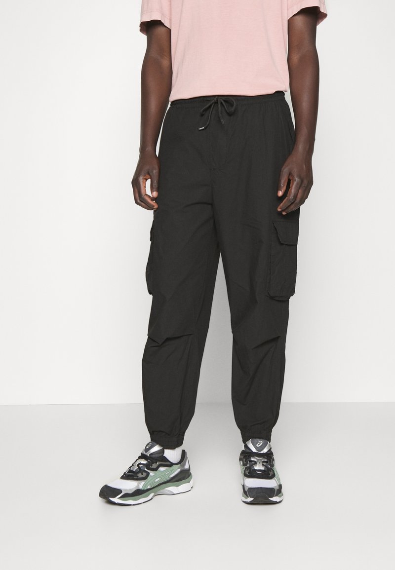 Homme portant un pantalon cargo noir avec taille à cordon et poignets élastiques, associé à des chaussures de sport blanches et noires.