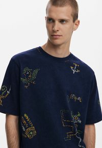 Marineblaues T-Shirt mit kurzen Ärmeln, das bunte bestickte Grafiken zeigt, darunter Vögel und abstrakte Formen, mit einem runden Ausschnitt und lockerer Passform.