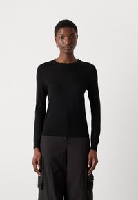 Μαύρο πλεκτό πουλόβερ με μακρύ μανίκι, στρογγυλή λαιμόκοψη, εφαρμοστή σιλουέτα και λεπτή υφή. Συνδυάζεται με σκούρες, φαρδιές cargo παντελόνες.