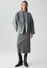 Graue Wolljacke im Oversize-Stil mit Revers, grauer Pullover darunter und grauer Midirock. Schwarze Knöchelstiefel runden das Outfit ab.