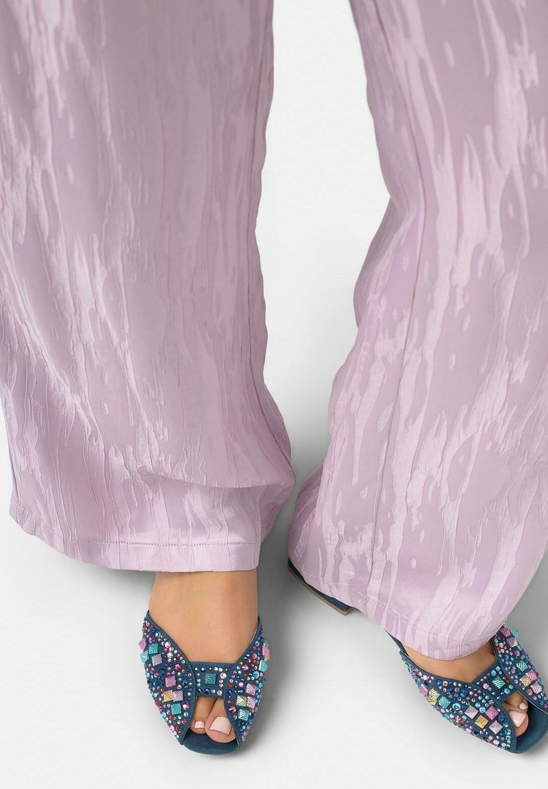 Pies con sandalias azules de punta abierta decoradas con brillantes cuadrados multicolores, combinadas con pantalones de pierna ancha de textura morado claro.