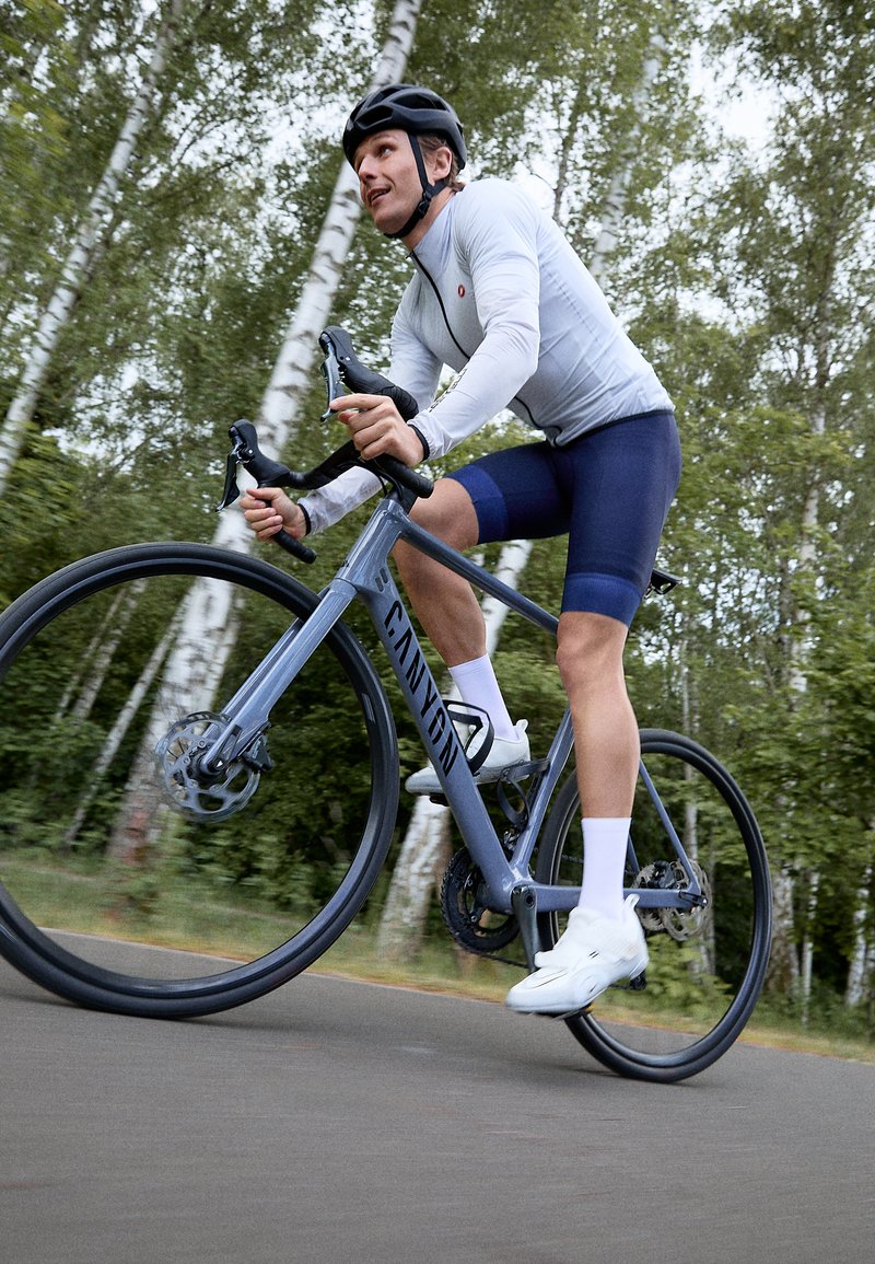 Vélo de route Canyon avec un cadre gris clair, des freins à disque et des pneus noirs. Cycliste en maillot à manches longues blanc et shorts bleus roulant sur un chemin.