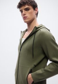 Sudadera con capucha y cremallera en verde oliva con textura acanalada, cordones negros y bolsillos laterales. Los materiales son suaves y el diseño es ajustado.