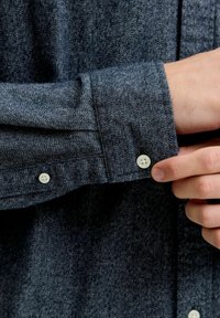 Roka balta pogu tumši pelēkās tekstūrētās garajās piedurknēs.