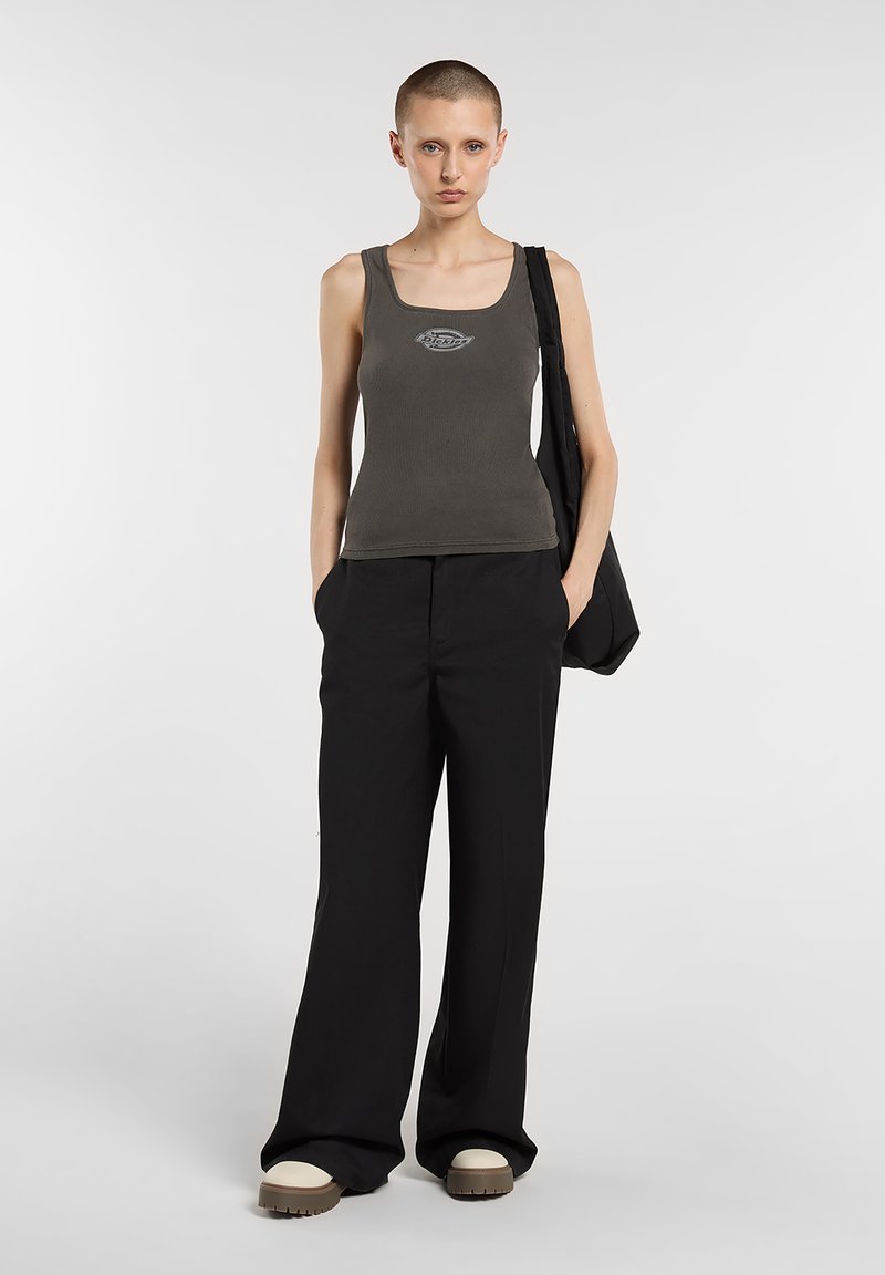 Graues, geripptes Tanktop mit Logo, kombiniert mit schwarzen, weit geschnittenen Hosen. Das Model hält eine schwarze Tasche und trägt cremefarbene und braune Schuhe.