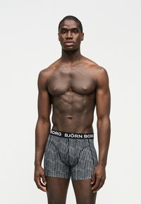 Ευχάριστος άνδρας με σκούρο δέρμα, όρθιος, φοράει ανθρακί εσώρουχα Björn Borg με ασπρόμαυρα σχέδια, ουδέτερη έκφραση προσώπου, απλό φωτεινό φόντο.