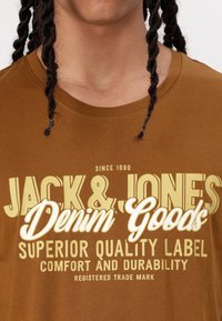 Camiseta de algodón marrón con estampado en blanco y amarillo, que incluye "JACK & JONES", "Denim Goods" y "SUPERIOR QUALITY LABEL". Diseño de cuello redondo.