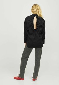 Maglietta nera con collo alto, dettaglio arricciato sul retro e scollatura a chiave; abbinata a pantaloni grigi e scarpe basse rosse.