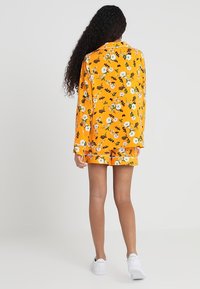 Gelbes Blumen-Pyjama-Set mit langärmligem Oberteil und Shorts mit weißen Details. Der Stoff hat eine weiche Textur und präsentiert weiße und grüne Blumen.