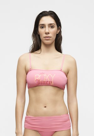 Fiatal nő hosszú sötét hajjal, rózsaszín bandeau bikini felsővel, melyen a „Roxy Juicy” felirat olvasható, és hozzáillő alsó résszel, fehér háttér előtt.