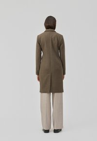 Olijfgroen wollen jas met een getailleerd ontwerp, lange mouwen, een geknipte kraag en knoopmanchetten, gecombineerd met lichtbeige wijde broekspijpen.