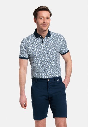 Polo à manches courtes blanc avec un motif géométrique bleu, col marine et poignets en marine. Porté avec un short marine, présentant un détail de bouton.