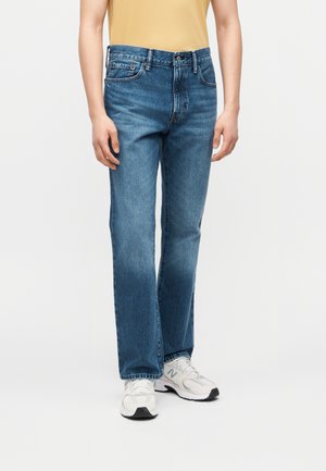 Osoba w niebieskich prostych dżinsach o średnim stanie, białych tenisówkach i włożonej do środka żółtej koszuli, stojąca na jednolitym tle.