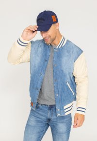 Veste bomber en denim avec un corps bleu clair, des manches crème, un col côtelé et des poignets rayés. Associée à un t-shirt gris et un jean bleu.