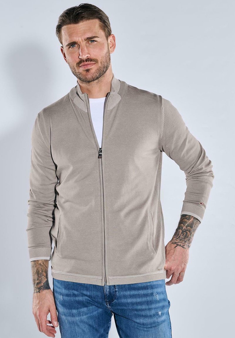 Helle beige Zip-Jacke aus glattem Stoff, mit einem gerippten Kragen, elastischen Bündchen und zwei Seitentaschen, getragen über einem weißen Hemd.