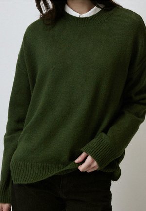 Personne portant un pull en maille vert foncé par-dessus une chemise blanche à col, tirant légèrement l'ourlet du pull avec la main droite.