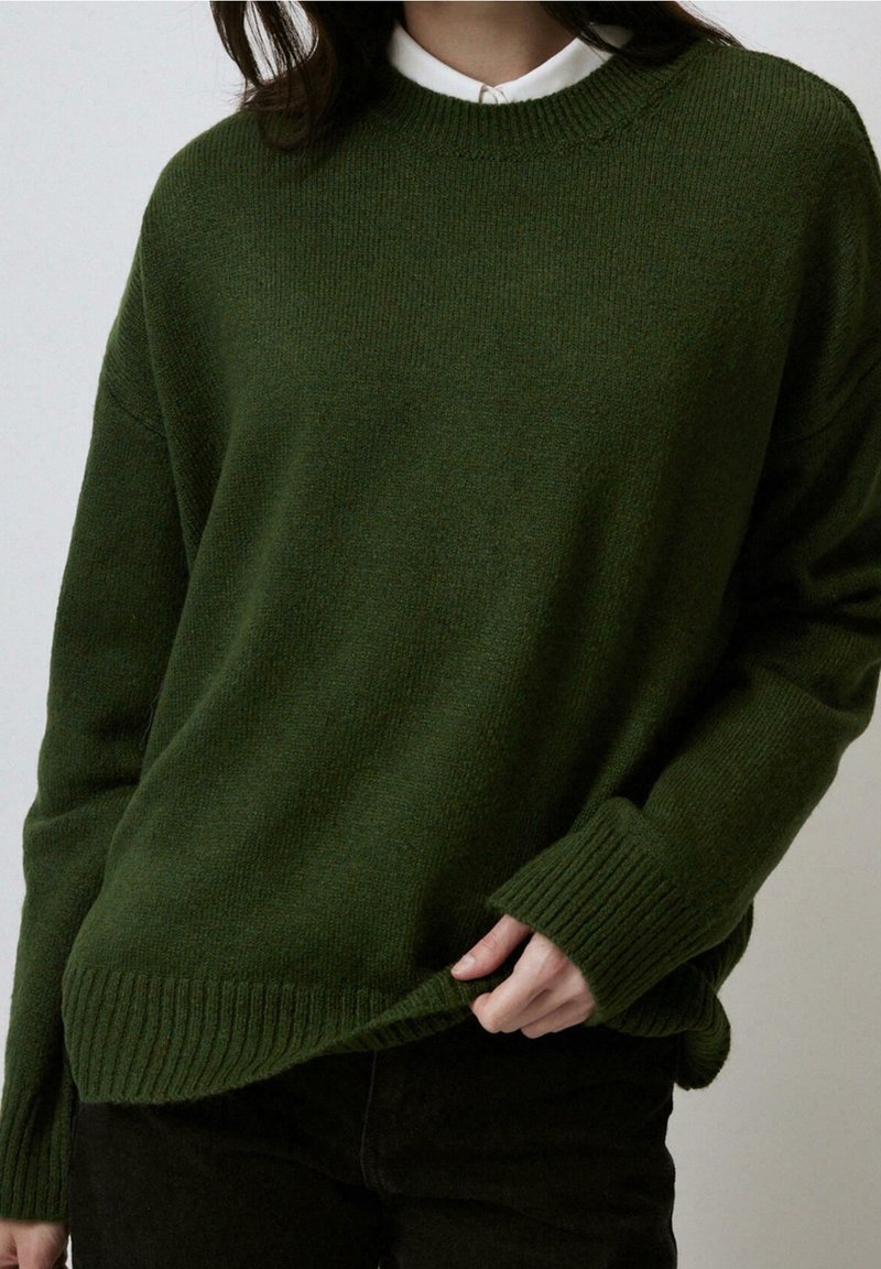 Personne portant un pull en maille vert foncé par-dessus une chemise blanche à col, tirant légèrement l'ourlet du pull avec la main droite.