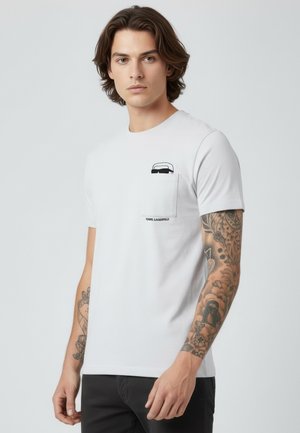 Junger Mann mit tätowierten Armen, der ein weißes Karl Lagerfeld T-Shirt mit einer kleinen Tasche und einem schwarzen Schriftzug darüber trägt.