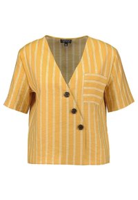 Camicia giallo senape a maniche corte con strisce verticali bianche, scollatura a V, taschino frontale e tre bottoni scuri sul lato sinistro.