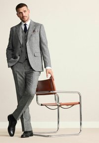 Grey textured suit with a tailored fit, complemented by a white shirt and dark tie. The model leans against a chrome and brown leather chair.