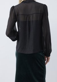 Blusa nera trasparente con righe verticali testurizzate, maniche a sbuffo e polsini con bottoni, abbinata a una gonna di velluto verde scuro.