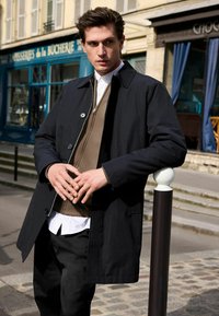 Palton negru cu guler, suprapus peste un pulover tricotat maro și o cămașă albă. Textură netedă și accente cu nasturi. Cadru urban casual.