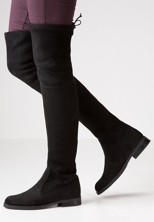 Legs wearing black suede over-the-knee flat boots with a back tie detail, paired with fitted maroon pants.