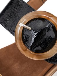 Detailní záběr lesklého černého pásku z lakované kůže s průhlednou hnědou kulatou přezkou a stříbrným zapínáním na trn.