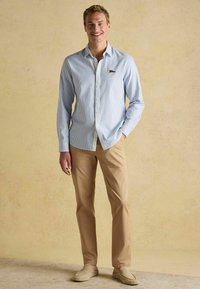 Jasnoniebieska koszula w prążki z długim rękawem i kołnierzykiem na guziki, beżowe spodnie oraz beżowe buty wsuwane, na neutralnym tle.