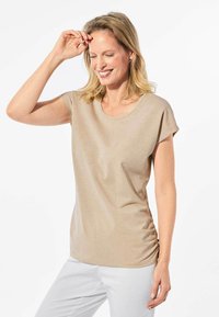 Beige Kurzarmoberteil mit Rundhalsausschnitt und seitlichem Raffungselement. Hergestellt aus einem glatten, leicht schimmernden Stoff, kombiniert mit weißen Hosen.