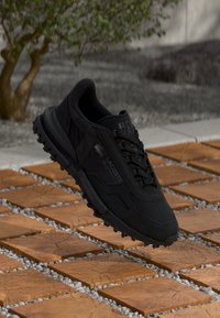 Adidași negri Lacoste cu un material superior texturat, închidere cu șireturi și talpă robustă din cauciuc, așezați pe un fundal din plăci de gresie.