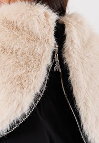 Colletto in pelliccia sintetica beige su tessuto nero con zip argentata visibile e uniche linguette in metallo a doppio cursore.