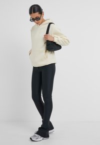Beige Kapuzenpullover mit einer Vordertasche, kombiniert mit schwarzen, figurbetonten Leggings. Das Modell trägt eine kleine schwarze Tasche und weiße Turnschuhe.