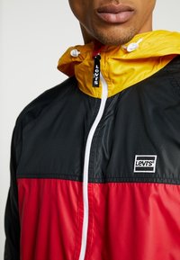 Mężczyzna w kurtce przeciwdeszczowej Levi's z żółtym kapturem, czarną górną częścią i czerwoną dolną, zapiętej pod szyję.