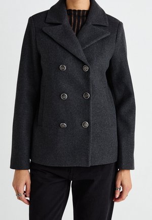 Blazer en laine gris foncé à double boutonnage avec six boutons, porté sur un top noir transparent rayé et un pantalon noir.