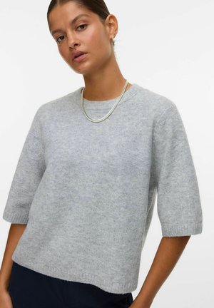 Vrouw in een lichtgrijze, korte mouw trui, met een zilveren ketting en donkere broek, staand tegen een eenvoudige witte achtergrond.