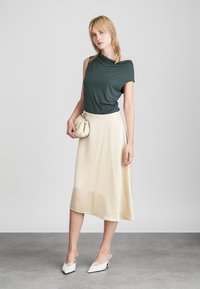 Sötétzöld, rövidujjú felső, amelyet egy krém színű, félhosszú selyemszerű szoknyával párosítanak. A modell egy bézs táskát tart, fehér magas sarkú papucsot visel.