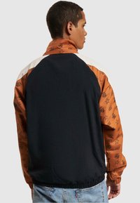 Sort og orange vindjakke med et mønstret ærme, der har stjerner og initialer; har høj krave og elastisk liv. 
