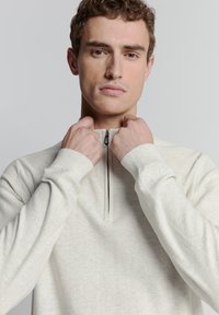 Lichtgrijs fleece pullover met een halve rits kraag, geribbelde manchetten en een gladde textuur. Het model past de kraag aan.