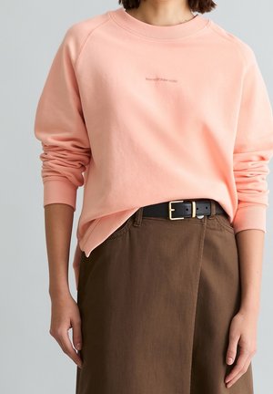 Femme portant un sweat-shirt pêche à manches longues avec les manches retroussées, rentrée dans une jupe marron taille haute et une ceinture noire avec une boucle dorée.