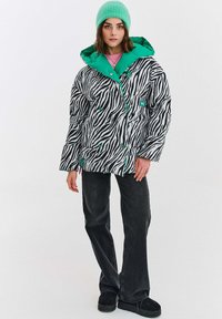 Žena oblečená v puffrovacej bunde so zebrovým vzorom a zeleným podšívaním, so zodpovedajúcou zelenou čiapkou, čiernymi džínami a čiernymi platformovými topánkami.