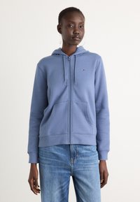 Hoodie bleu zippé en tissu doux, avec une poche avant et un petit logo sur la poitrine. Associé à un jean bleu clair.